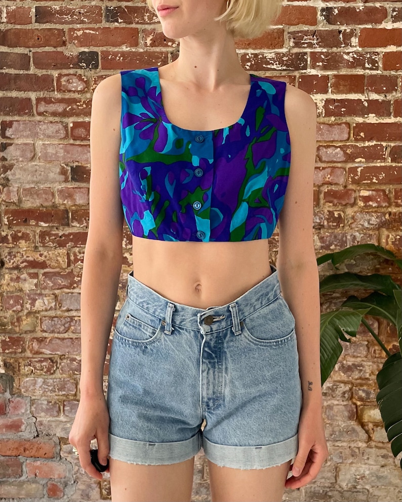 May include: A sleeveless crop top with a colourful abstract floral print in shades of blue, green, and purple. The top has a button closure at the front.