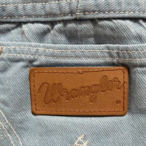 Puede incluir: Primer plano de tela vaquera azul claro con costuras blancas. Un parche de cuero marr&oacute;n con la palabra "Wrangler" en relieve est&aacute; cosido en el bolsillo de mezclilla. El logotipo de Wrangler es visible.