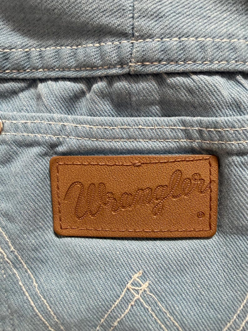 Puede incluir: Primer plano de tela vaquera azul claro con costuras blancas. Un parche de cuero marr&oacute;n con la palabra "Wrangler" en relieve est&aacute; cosido en el bolsillo de mezclilla. El logotipo de Wrangler es visible.