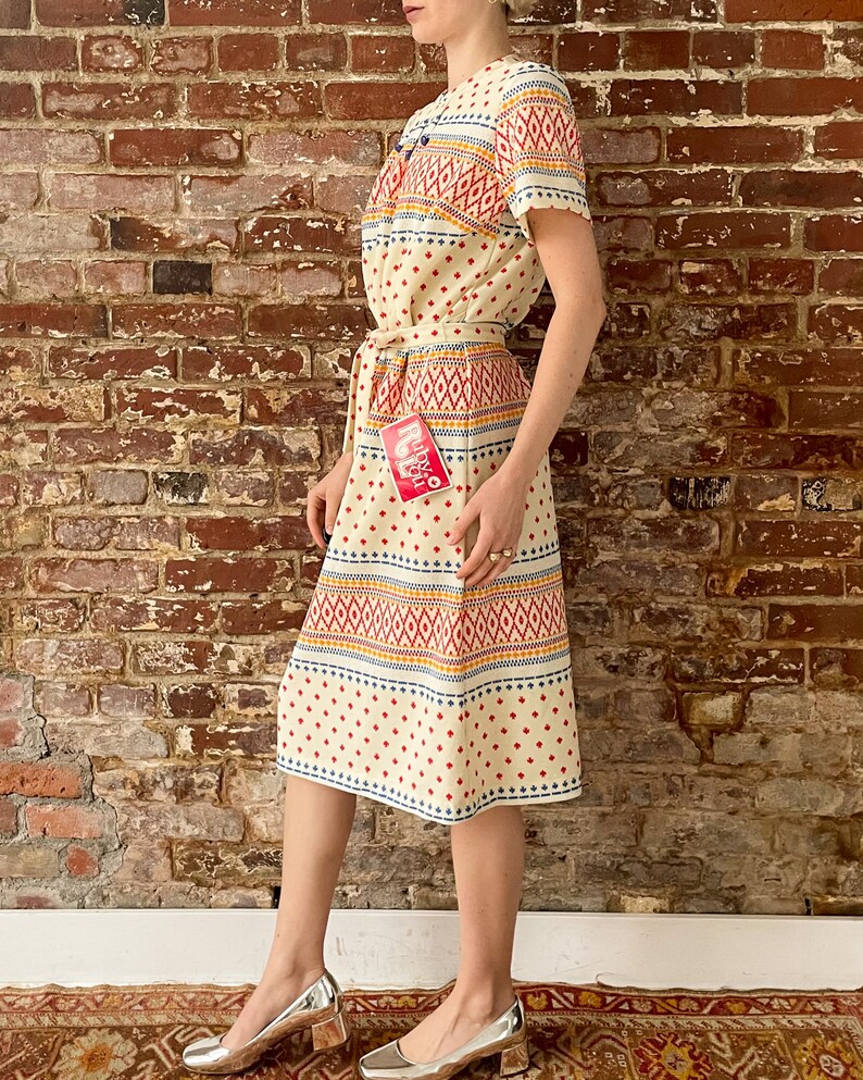 May include: A cream-colored dress with a geometric pattern in red, blue, and yellow. The dress has a short sleeve and a tie belt. The dress has a tag that says "Royal" in red letters.