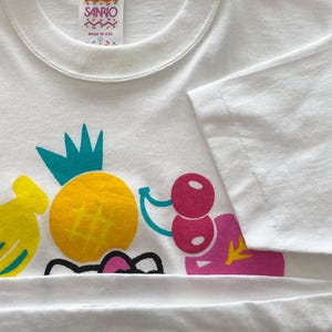 Puede incluir: Camiseta blanca con un dise&ntilde;o de frutas colorido. El dise&ntilde;o incluye un pl&aacute;tano amarillo, una pi&ntilde;a amarilla con una corona turquesa y cerezas rosas. La camiseta tiene una etiqueta Sanrio y est&aacute; hecha en los EE. UU.