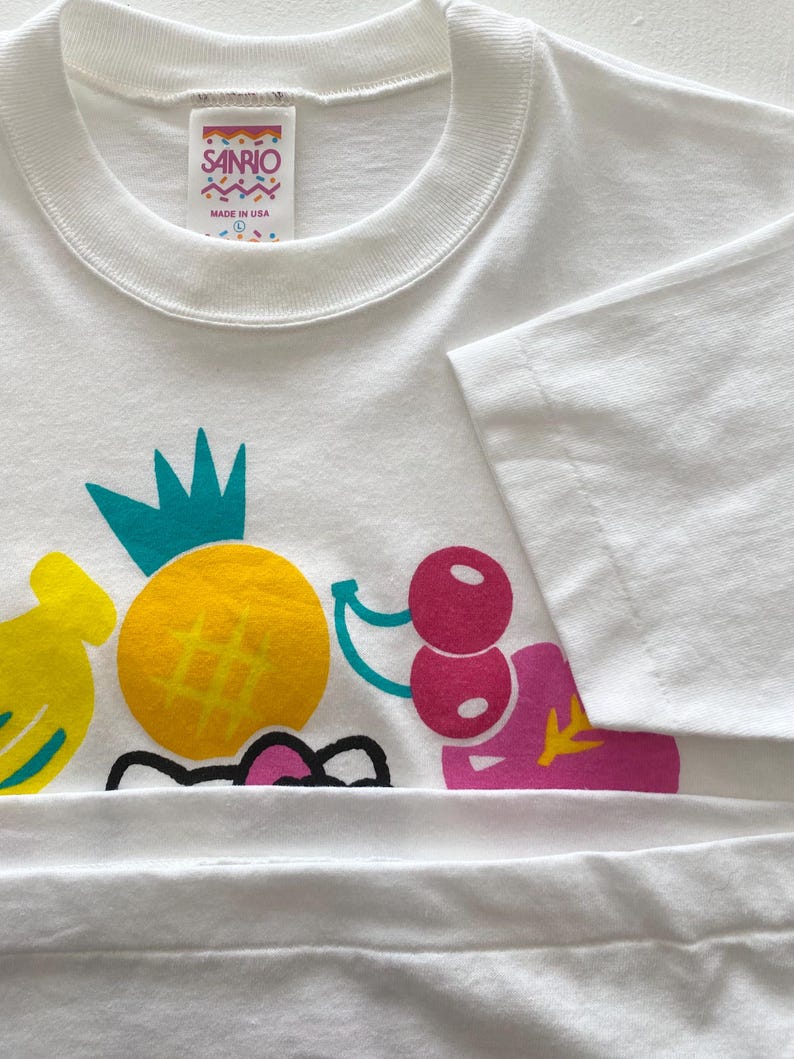Puede incluir: Camiseta blanca con un dise&ntilde;o de frutas colorido. El dise&ntilde;o incluye un pl&aacute;tano amarillo, una pi&ntilde;a amarilla con una corona turquesa y cerezas rosas. La camiseta tiene una etiqueta Sanrio y est&aacute; hecha en los EE. UU.