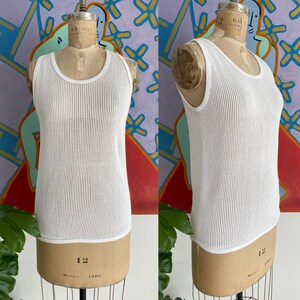 May include: A white, sleeveless tank top with a textured, open-knit design. The top has a rounded neckline and a white trim along the edges. The garment is displayed on a vintage mannequin.