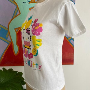 Puede incluir: Camiseta blanca con un gr&aacute;fico colorido de Hello Kitty rodeada de frutas y flores. El dise&ntilde;o incluye elementos rosas, amarillos y verdes, con el texto "Hello Kitty" en rosa. La camiseta se muestra en un maniqu&iacute;.
