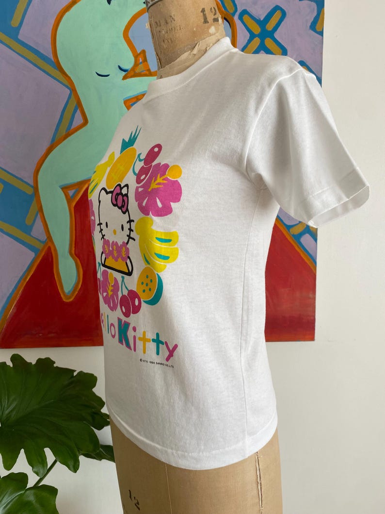 Puede incluir: Camiseta blanca con un gr&aacute;fico colorido de Hello Kitty rodeada de frutas y flores. El dise&ntilde;o incluye elementos rosas, amarillos y verdes, con el texto "Hello Kitty" en rosa. La camiseta se muestra en un maniqu&iacute;.