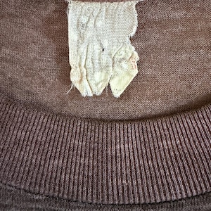 May include: Close-up of a brown t-shirt showing a ribbed collar and a torn, off-white label. The label is made of a woven material and has a few small holes. The shirt's fabric has a slightly textured appearance.