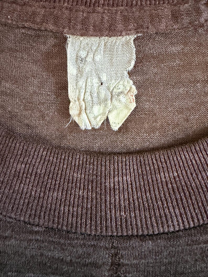 May include: Close-up of a brown t-shirt showing a ribbed collar and a torn, off-white label. The label is made of a woven material and has a few small holes. The shirt's fabric has a slightly textured appearance.