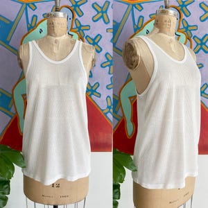 May include: White mesh tank top with a scoop neck and a slightly rounded hem. The tank top is displayed on a vintage mannequin. The fabric has a perforated texture. The mannequin is on a neutral background.