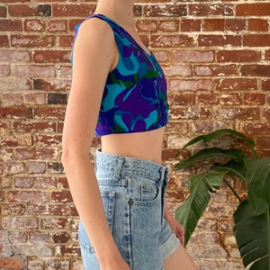 Peut inclure: Un haut court sans manches avec un imprim&eacute; abstrait bleu, violet et vert. Le haut a une fermeture boutonn&eacute;e &agrave; l'avant. Le mannequin porte le haut avec un short en jean bleu clair.