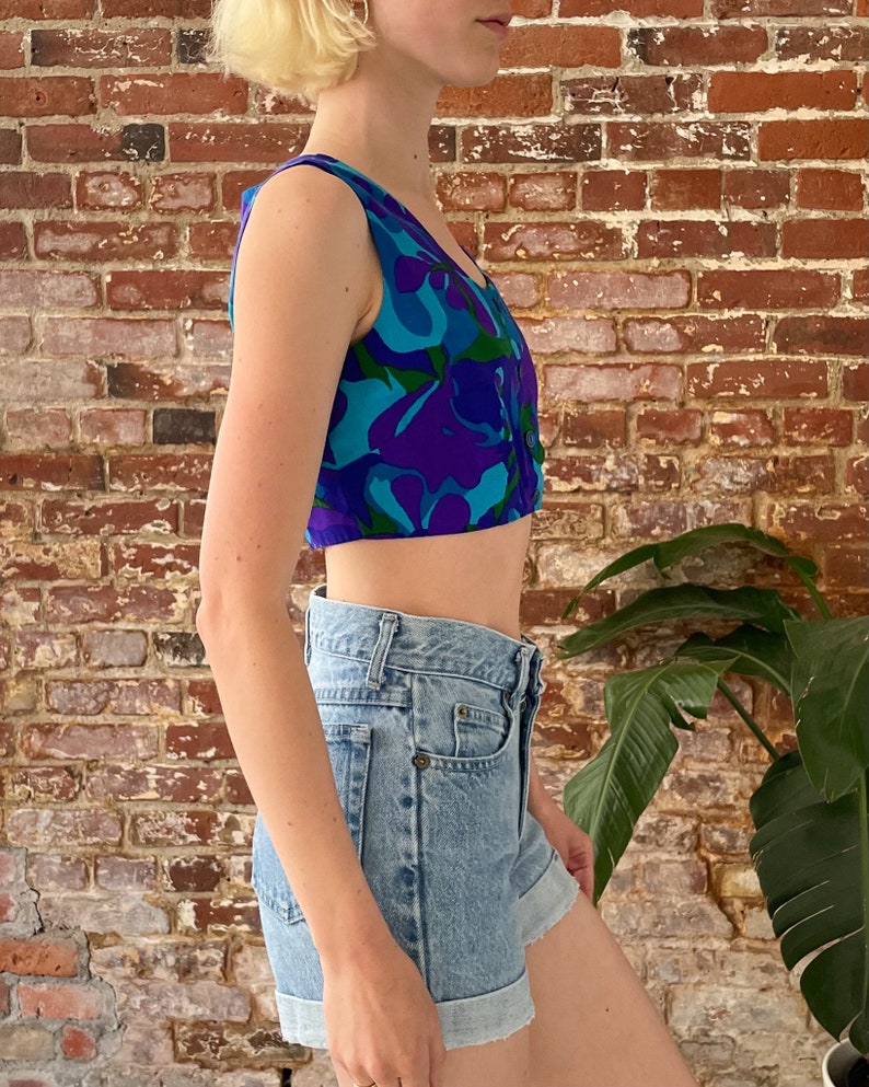 Peut inclure: Un haut court sans manches avec un imprim&eacute; abstrait bleu, violet et vert. Le haut a une fermeture boutonn&eacute;e &agrave; l'avant. Le mannequin porte le haut avec un short en jean bleu clair.