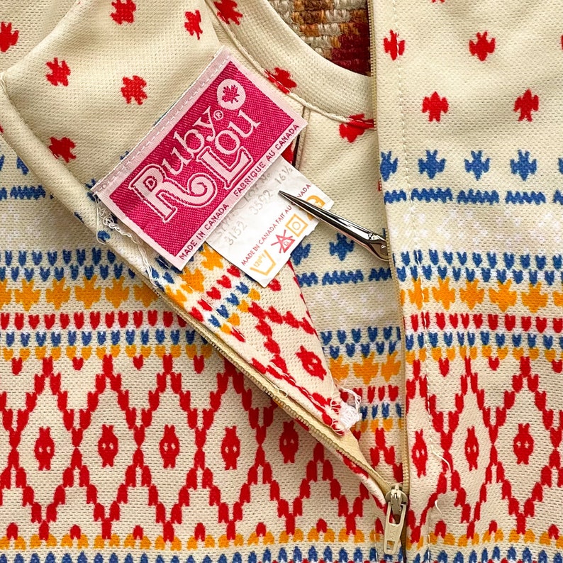 May include: A white zippered sweater with a red, yellow, and blue geometric pattern. The sweater has a pink label that reads "Ruby Lou" and "Made in Canada Fabrique au Canada".