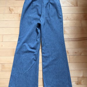 Puede incluir: Pantalones vaqueros azul claro con dise&ntilde;o de pernera ancha. Los pantalones est&aacute;n colocados planos sobre una superficie de madera, mostrando la parte trasera de la prenda. La cintura presenta pliegues y cierre de bot&oacute;n. La tela parece ser de mezclilla de peso medio.
