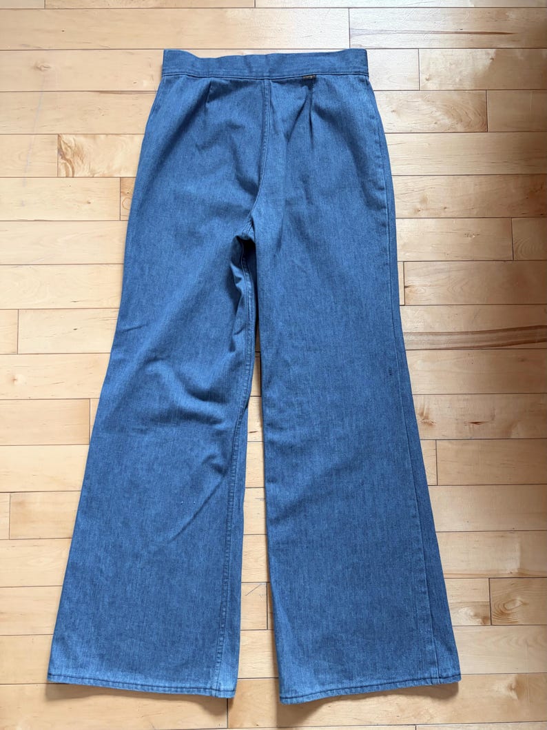 Puede incluir: Pantalones vaqueros azul claro con dise&ntilde;o de pernera ancha. Los pantalones est&aacute;n colocados planos sobre una superficie de madera, mostrando la parte trasera de la prenda. La cintura presenta pliegues y cierre de bot&oacute;n. La tela parece ser de mezclilla de peso medio.