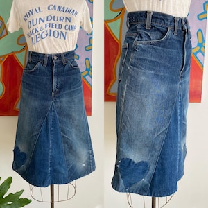 May include: A vintage denim skirt with a heart-shaped patch. The skirt is a medium wash blue with a high waist and an A-line silhouette. The skirt has a heart-shaped patch on the front. The skirt is displayed on a mannequin.