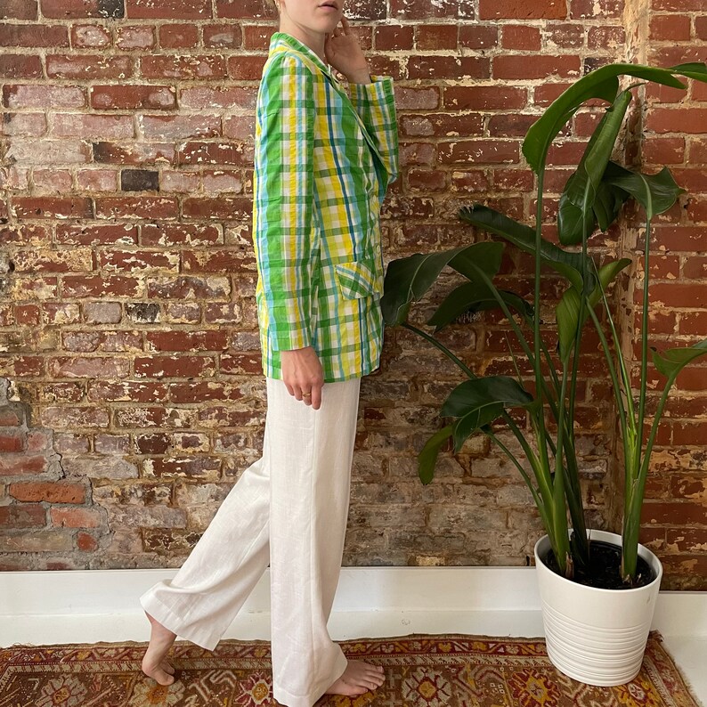 May include: A person wearing a green and yellow plaid blazer with white trousers. The blazer has a button fastening and a pocket on the left side. The person is standing in front of a brick wall with a green plant in a white pot.