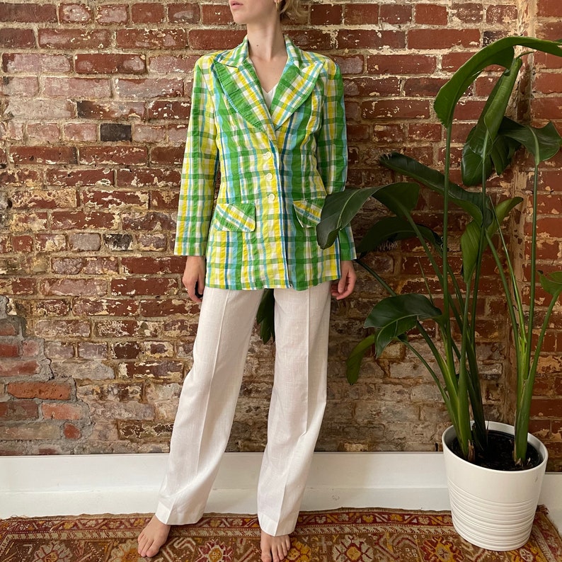 May include: A green and yellow plaid blazer with a double-breasted closure and a white collared shirt underneath. The blazer is worn with white wide-leg trousers.