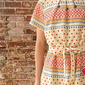 May include: A white dress with a geometric pattern in red, blue, and yellow. The dress has a short sleeve and a tie belt.