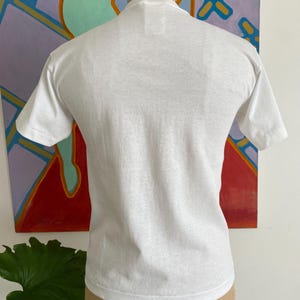 Puede incluir: Una camiseta blanca lisa de manga corta. La prenda se exhibe en un maniqu&iacute;. El fondo presenta una pintura abstracta colorida con tonos rojos, azules y naranjas. La tela de la camiseta parece ser de algod&oacute;n ligero.
