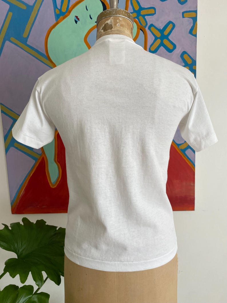 Puede incluir: Una camiseta blanca lisa de manga corta. La prenda se exhibe en un maniqu&iacute;. El fondo presenta una pintura abstracta colorida con tonos rojos, azules y naranjas. La tela de la camiseta parece ser de algod&oacute;n ligero.