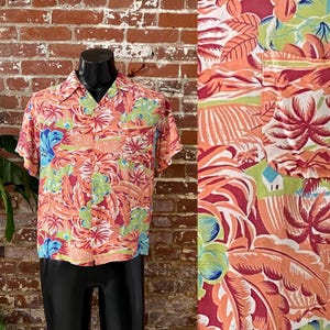 May include: A short-sleeved button-up shirt with a tropical print featuring palm trees, flowers, and a small house. The shirt is made of a lightweight fabric and has a relaxed fit.