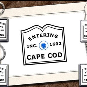 May include: Keychains and a necklace featuring a square pendant with the text "ENTERING CAPE COD INC. 1602" in a black and white design. The pendants have a silver border and are displayed on a brown and white background.
