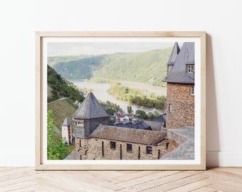 May include: A scenic view from a castle overlooking a river winding through a valley. The castle is made of stone and has a pointed roof. The river is surrounded by lush green trees and hills.