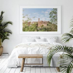 Op de afbeelding: Een ingelijste landschapsfoto van een Europese stad met een hoge klokkentoren, boven een bed met wit beddengoed. De kamer is ingericht met planten in manden en potten, en een houten bank aan het voeteneinde van het bed.