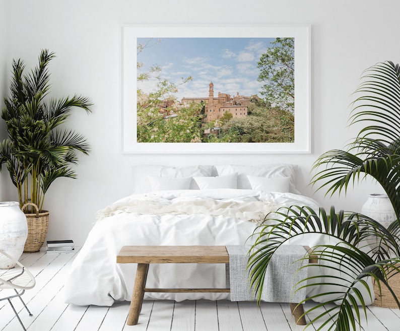 Op de afbeelding: Een ingelijste landschapsfoto van een Europese stad met een hoge klokkentoren, boven een bed met wit beddengoed. De kamer is ingericht met planten in manden en potten, en een houten bank aan het voeteneinde van het bed.