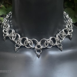 May include: A silver-colored chainmail choker necklace with a spiked design. The necklace is made of interlocking rings and features several downward-facing spikes. The text "Crystal Armoury" is visible at the bottom of the image.