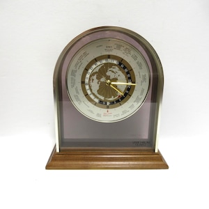May include: A mantel clock with a wooden base and an arched glass frame. The clock face features a world map, a 24-hour rotating ring, and a list of global cities. Gold-colored hands indicate the time, and the text Vacheron Quartz is visible.