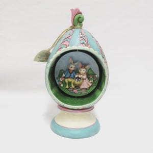 May include: An Easter-themed decorative egg ornament. The egg is light blue with pink accents and a green interior. Inside, two bunny figures are depicted. The ornament sits on a white and blue base and is topped with a pink flower.