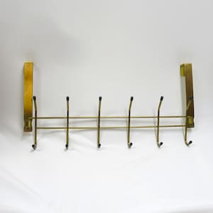 May include: A gold-toned metal over-the-door coat rack featuring six double hooks. Each hook is tipped with a small black protective cap. The rack is constructed from thin wire bars and includes two flat vertical brackets for mounting.