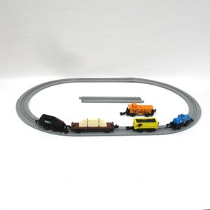 May include: A toy train set featuring a black locomotive, a flatcar with white cargo, a yellow hopper car, and a blue tank car on an oval gray plastic track. An orange caboose with Illinois Central text sits inside the track loop.