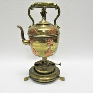 May include: Antique brass and copper kettle with a decorative design and a stand with a burner. The kettle has a spout and a handle.