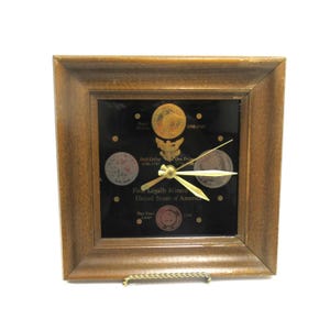 May include: A square wall clock with a medium-brown wooden frame and a black face. The clock features gold-colored hands and displays several historical United States coins with text reading First Legally Minted United States of America.