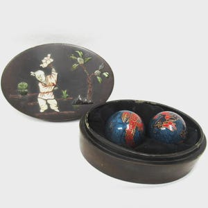 May include: A set of two blue decorative Chinese exercise balls with red and gold designs, nestled in a black velvet-lined wooden case. The lid features an inlaid scene of a child and a tree.