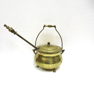 May include: A vintage brass pot with a lid, handle, and three small feet. A long brass spoon rests inside the pot. The pot has a rounded shape and a tarnished finish, suggesting age. The handle is arched and attached to the pot.