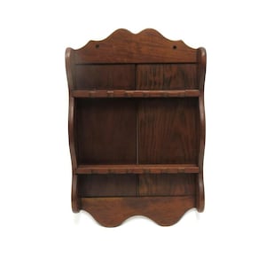 May include: A dark brown wooden wall shelf with two shelves and a decorative scalloped top and bottom edge. The shelf has a curved design on the sides and is made of solid wood.