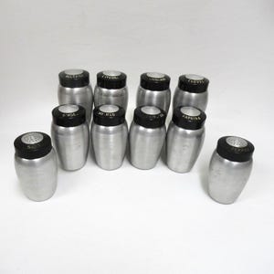 May include: A collection of vintage spice shakers with silver-colored bodies and black lids. The lids are labeled with spice names like "Salt," "Pepper," "Ginger," and "Cinnamon." The shakers are arranged on a white surface.