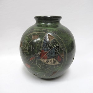 May include: A round, dark green vase featuring an etched design of a hummingbird in flight. The bird is detailed with red, blue, and white accents, surrounded by engraved foliage patterns. The vase has a smooth, glossy finish and a narrow neck opening.