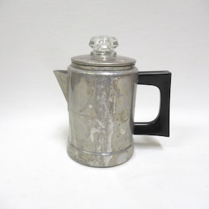 May include: Vintage silver-toned coffee percolator with a clear glass knob on the lid and a black handle. The metal pot has a spout and a cylindrical body. A classic kitchenware item.