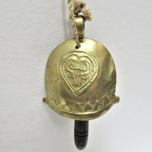 May include: A brass bell with a heart-shaped design featuring a bull's head. The bell is attached to a brown rope and has a wooden handle.