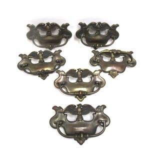 May include: A set of five matching brass drawer pulls, each featuring a central eagle motif at the top and a curved, swinging handle attached to a decorative backplate. The metal has a darkened, aged patina with visible highlights on the eagle.