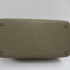 May include: A rectangular, tan-colored automotive armrest with a textured surface. The armrest has rounded corners and four circular mounting points, one in each corner. The item is likely a replacement part.