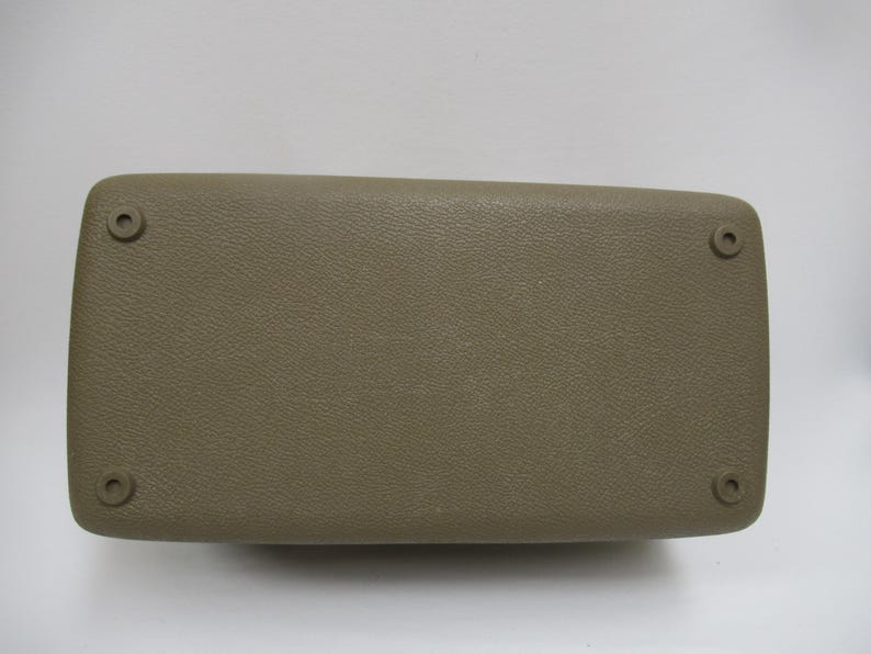 May include: A rectangular, tan-colored automotive armrest with a textured surface. The armrest has rounded corners and four circular mounting points, one in each corner. The item is likely a replacement part.