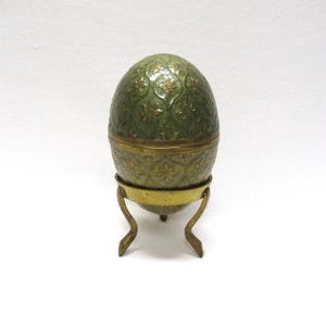 May include: A decorative egg-shaped container with a green, floral-patterned exterior and brass accents. The egg sits on a brass stand with curved legs, creating an elegant display piece. The container is likely used for jewelry or small keepsakes.