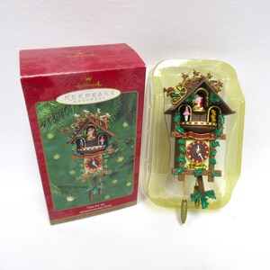 May include: A Hallmark Keepsake cuckoo clock ornament in a red box. The brown wooden-style clock features a Santa figure, a small girl, a teddy bear, green holly leaves, a red clock face with white hands, and two hanging pinecone-shaped weights.