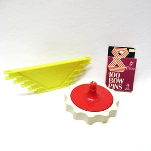 May include: A set of three yellow and white plastic tools for making bows. The tools include a bow maker, a bow pin holder, and a bow pin measuring tool. The bow pin holder has a red top and says "Bow Maker" on the side. The bow pin measuring tool has the words "Bow Maker" printed on it. The bow pin holder has a package of 100 bow pins next to it. The package is purple and white and says "100 Bow Pins" and "Pixie" on it.