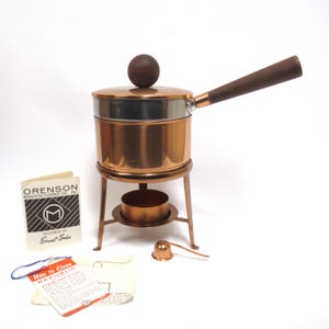 May include: A copper fondue pot with a stainless steel rim, a dark wooden handle, and a matching wooden knob on the lid. It sits on a copper stand with a burner cup and a small copper snuffer. Includes a booklet labeled Orenson Manufacturing Co., Inc.