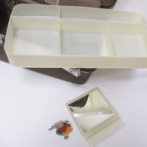 May include: A beige plastic organizer tray with multiple compartments, a small mirror, and a key with a colorful charm. The tray is part of a larger brown case, likely for cosmetics or personal items.